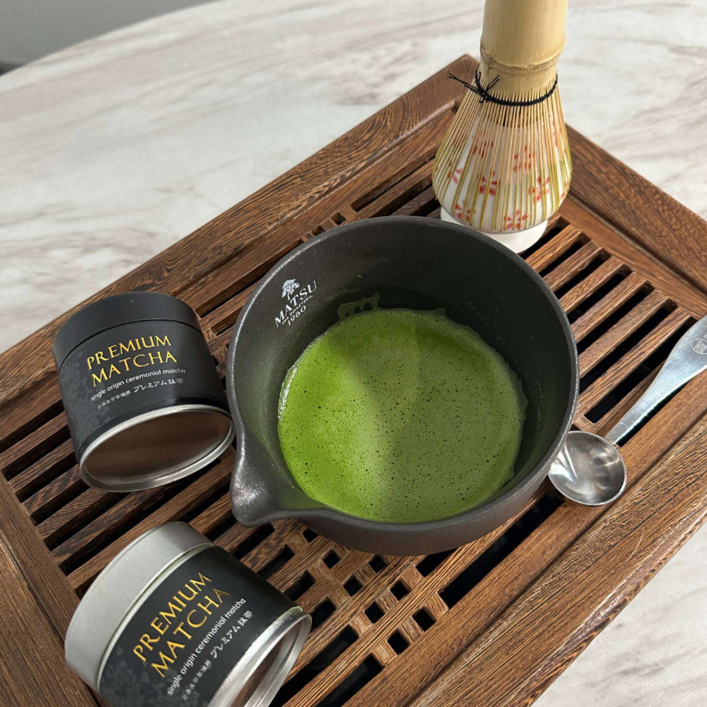 Matcha green tea set with a bowl of matcha, containers labeled 'Premium Matcha', and a whisk on a wooden tray.